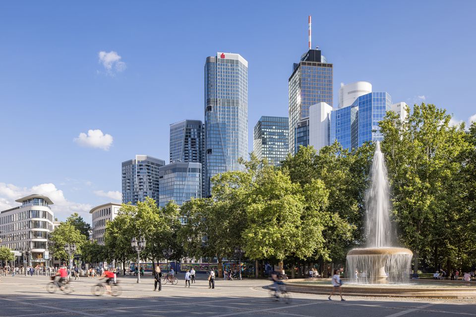 Außenaufnahme eines städtischen Platzes mit großem Brunnen, umgeben von dicht stehenden Hochhäusern und baumgesäumten Wegeflächen im Frankfurter Bankenviertel.