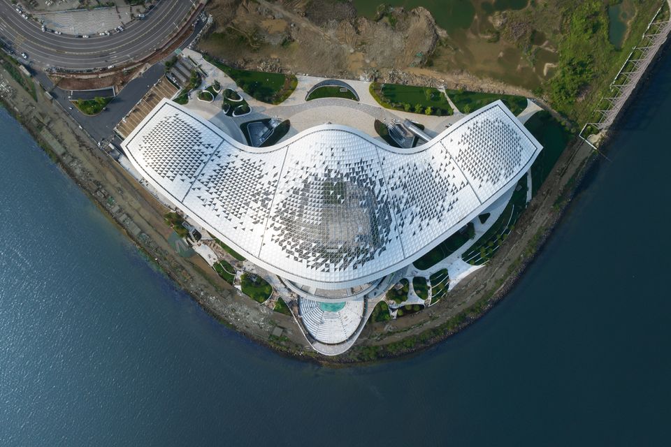 Draufsicht auf ein großflächiges Kulturgebäude mit geschwungener Dachform, Musterung aus polygonalen Paneelen und umlaufenden Außenbereichen am Wasser.