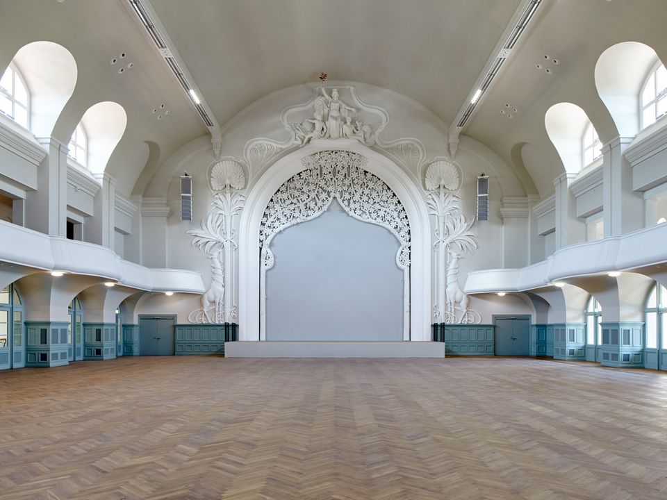 Innenaufnahme eines großen Saalraums mit kunstvoll ausgearbeitetem Bühnenportal, seitlichen Emporen und hellem Deckengewölbe, die die historische Raumgestaltung hervorheben.