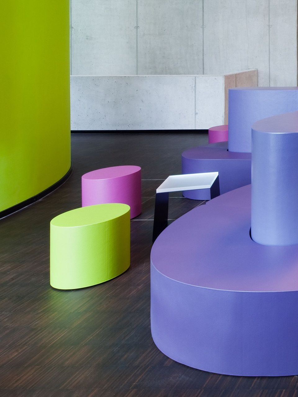 Innenraum mit farbigen Sitzmodulen in organischen Formen vor Betonwand, modernes Design mit kräftigen Grün-, Pink- und Violetttönen