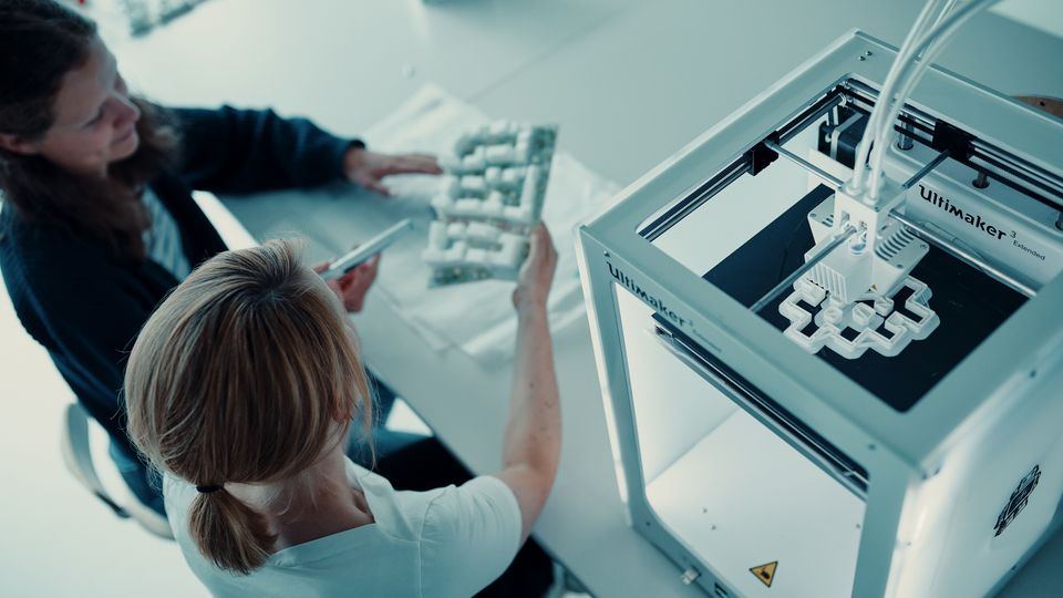 Blick auf einen 3D-Drucker während der Fertigung eines Modells. Personen betrachten ein gedrucktes Bauteil.