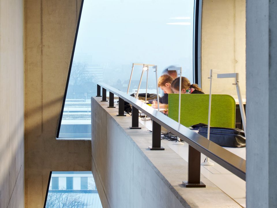 Lese- und Arbeitsbereich in moderner Bibliothek mit weißen Regalen und grünen Sichtschutzpaneelen vor Betonwand
