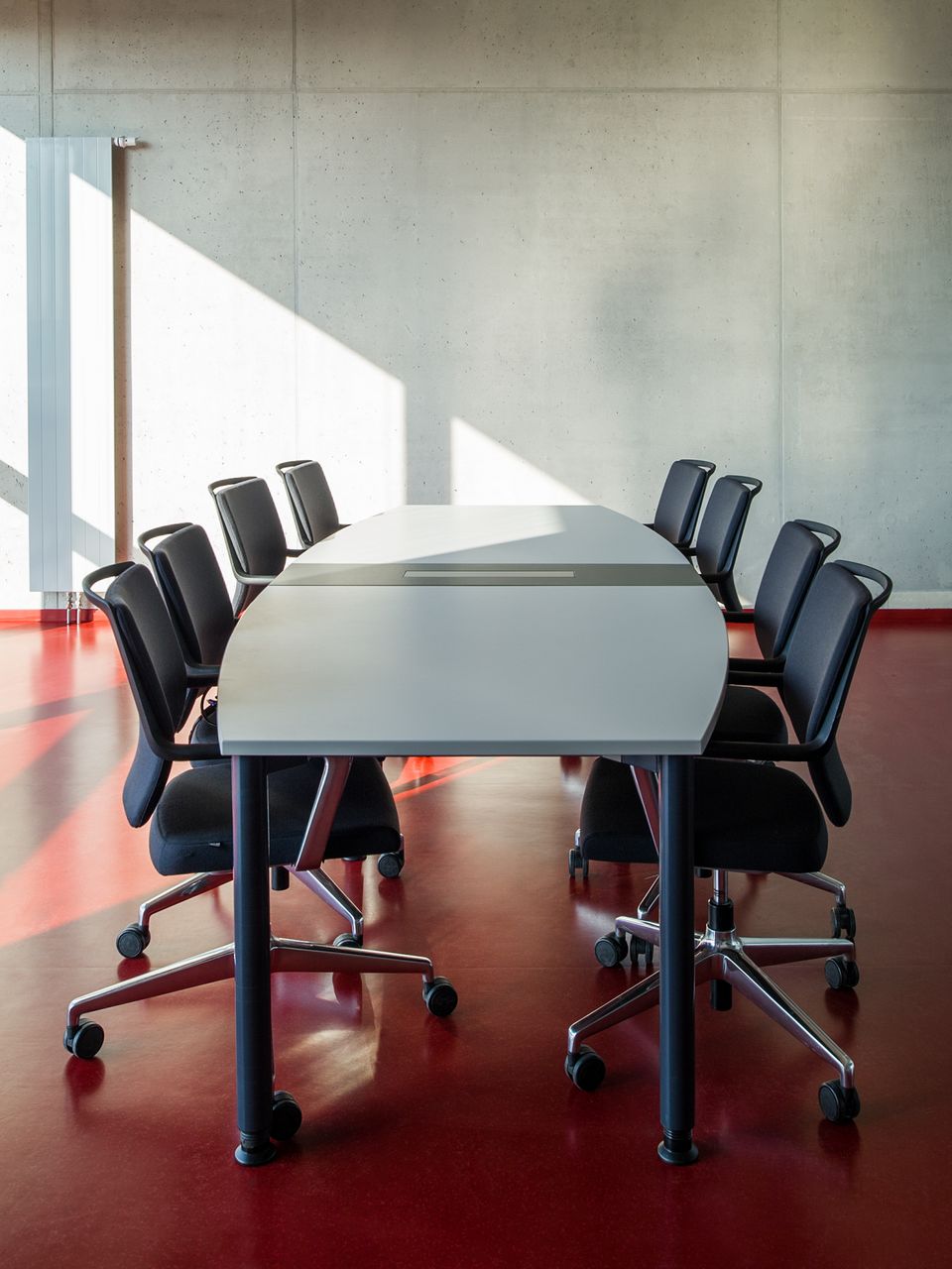 Innenaufnahme eines Seminarraums mit langem Arbeitstisch, flexiblen Bürostühlen und glatter Sichtbetonwand.