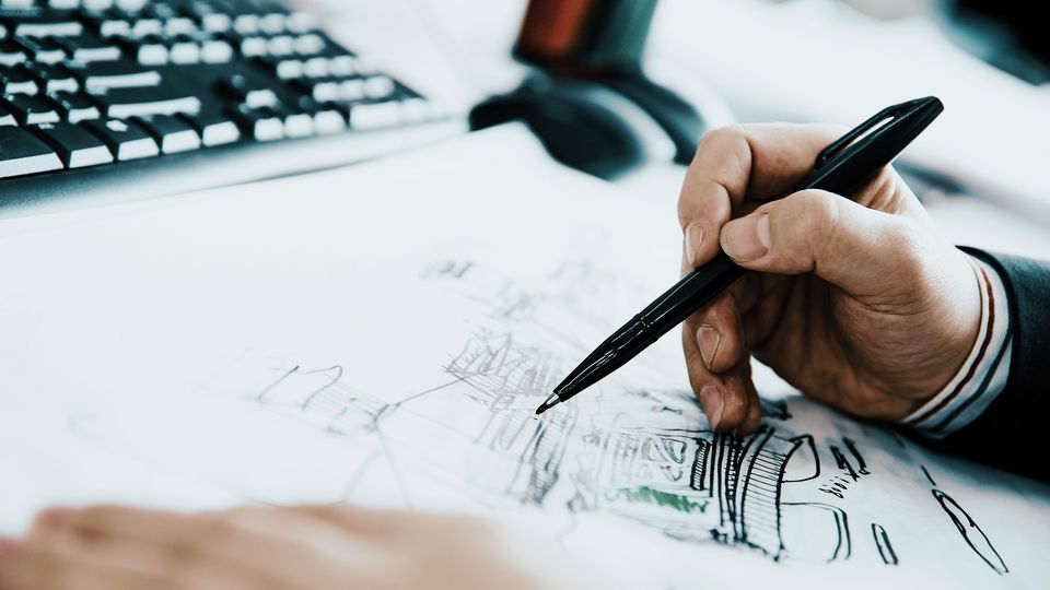Detailaufnahme einer Hand, die mit einem Stift an einer architektonischen Zeichnung arbeitet, im Umfeld eines Arbeitsplatzes mit Tastatur und Monitor.