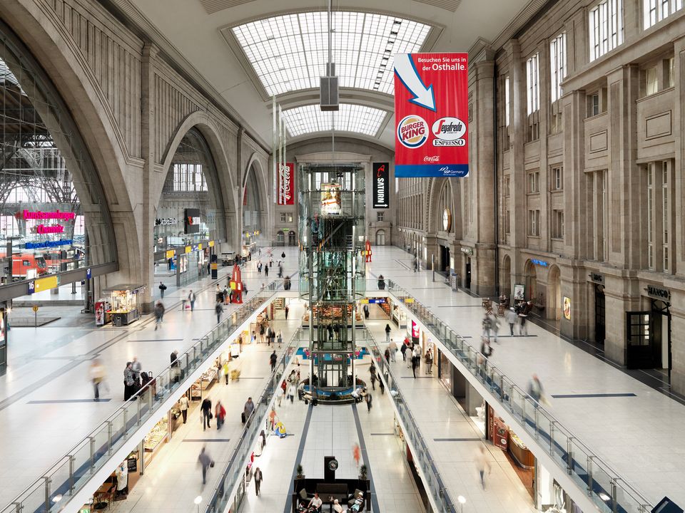 Innenaufnahme der zentralen Bahnhofshalle mit mehrgeschossigen Ladenebenen, verglastem Aufzugskern, breiten Erschließungswegen und raumprägender Gewölbestruktur.