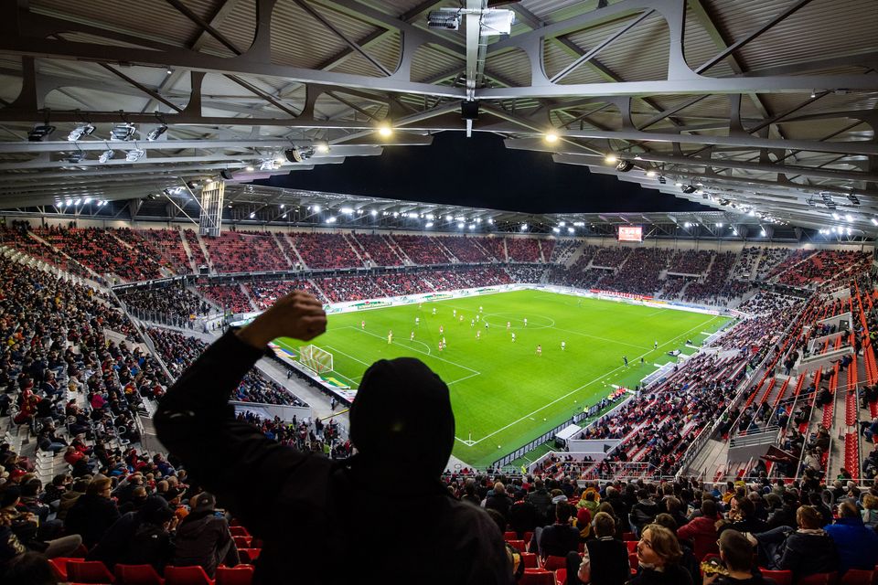 Blick aus der Kurve auf das Spielfeld im beleuchteten Stadion, rote Sitzreihen und dynamische Dachkonstruktion, volle Tribünen