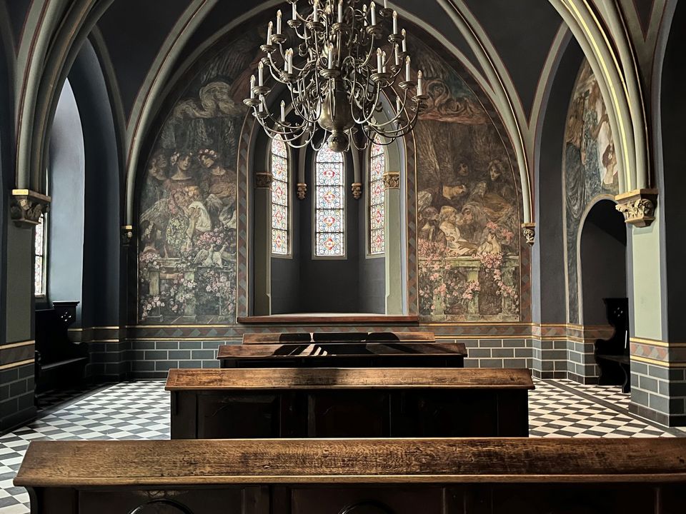 Innenaufnahme einer Kapelle mit gewölbter Architektur, großformatigen Wandmalereien, ornamentierten Fenstern und traditionellen Holzbänken im vorderen Bereich.