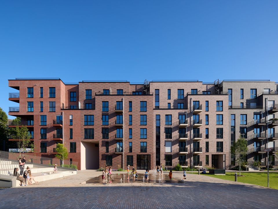 Außenaufnahme eines modernen Wohngebäudes mit differenzierten Klinkerfassaden, Balkonen und einem belebten Vorplatz mit Wasserspiel in einem urbanen Quartier.