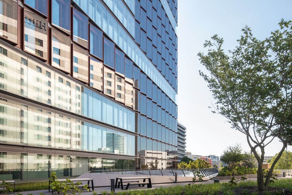 Terrassenbereich mit Sitzbänken vor Glasfassade eines Bürogebäudes, umgeben von Grünflächen und Bäumen