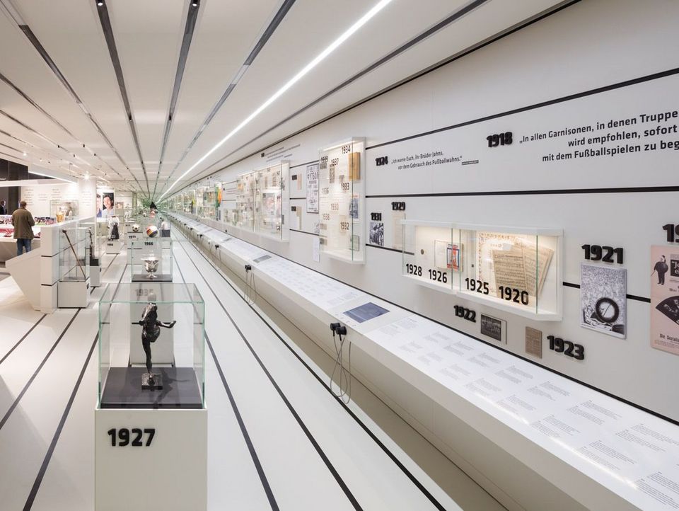 Innenaufnahme eines Museums mit einer langgestreckten Vitrinenwand, historischen Exponaten, Jahresmarkierungen und einer chronologisch aufgebauten Präsentation zur Fußballgeschichte.