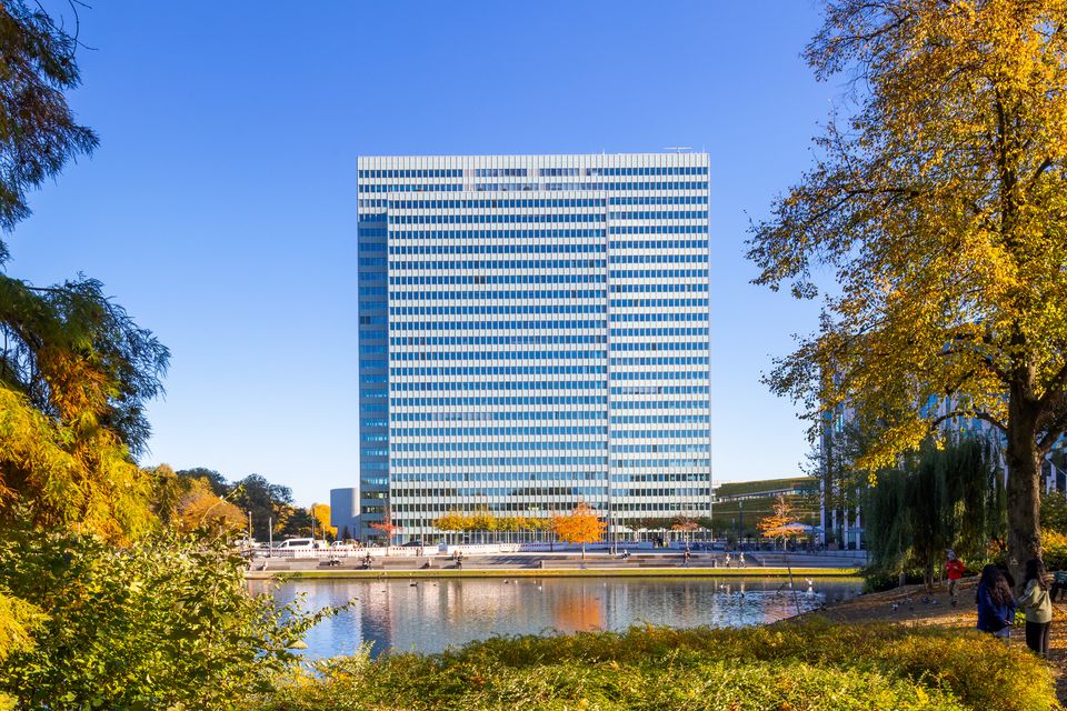 Frontansicht eines Hochhauses mit Glasfassade, eingebettet in Parklandschaft mit Wasserfläche und herbstlicher Vegetation