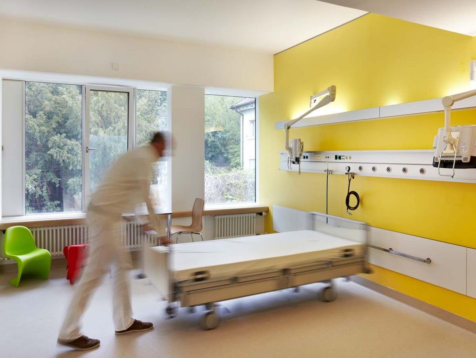 Patientenzimmer mit gelber Akzentwand, großem Fenster und moderner Ausstattung, funktionales Design mit klarer Struktur