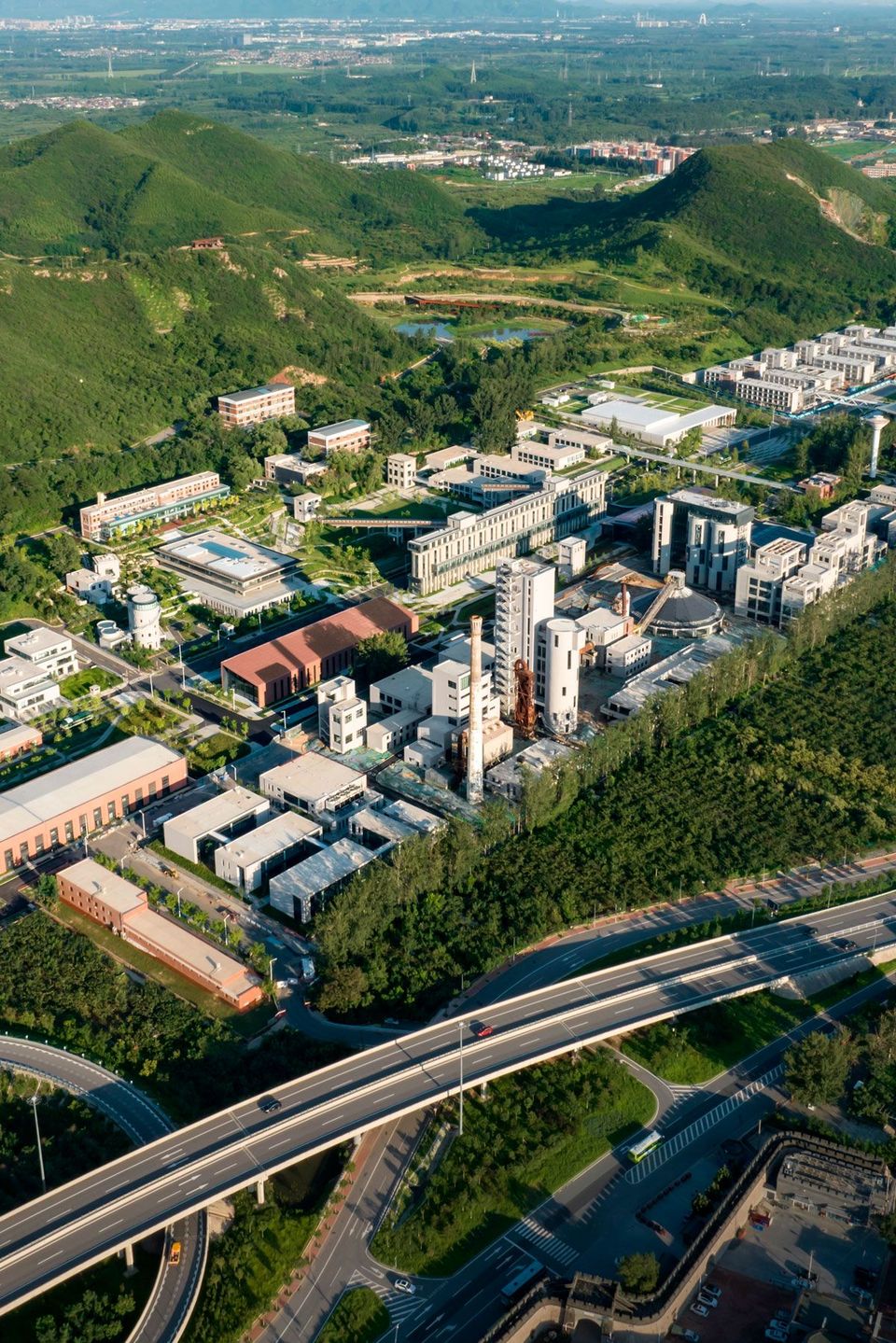Luftaufnahme eines Industriecampus mit Silos, Produktionshallen und begrünten Flächen, umgeben von Bergen