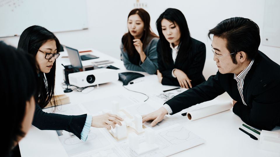 Innenaufnahme eines Besprechungsraums mit mehreren Personen an einem Tisch, die gemeinsam an einem Architekturmodell und technischen Plänen arbeiten.