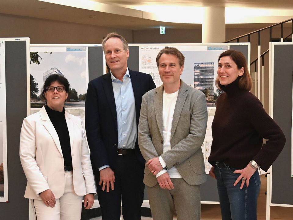 Gruppenaufnahme vor mehreren Ausstellungstafeln in einem Innenraum, umgeben von Präsentationsflächen und architektonischen Visualisierungen im Hintergrund.