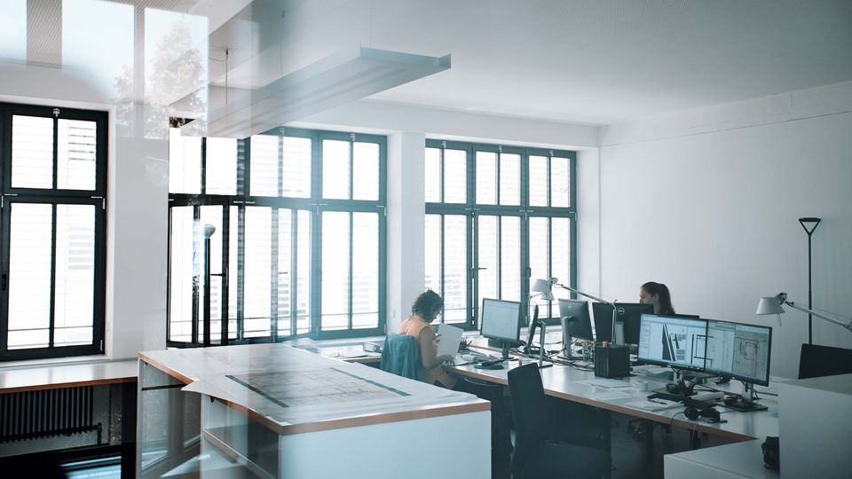 Innenaufnahme eines hellen Büroarbeitsbereichs mit großen Fensterflächen, mehreren Arbeitsplätzen, Monitoren und technischen Zeichentools.
