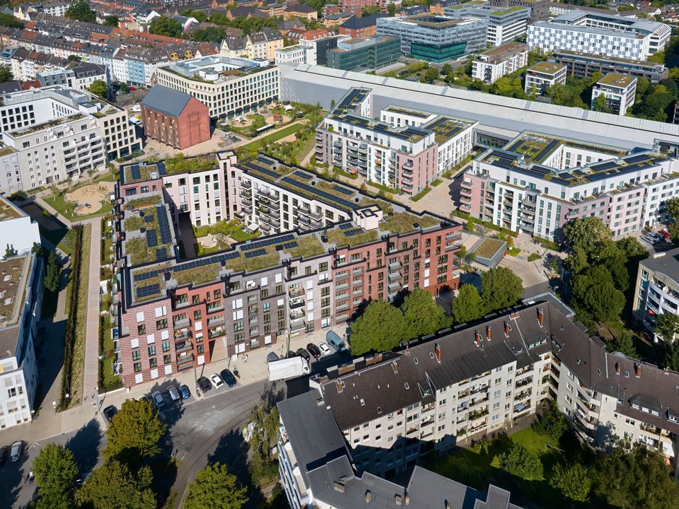 Luftaufnahme eines modernen Wohnensembles mit abwechslungsreicher Dachlandschaft, begrünten Terrassen und klar gegliederter Quartiersstruktur im städtischen Umfeld.