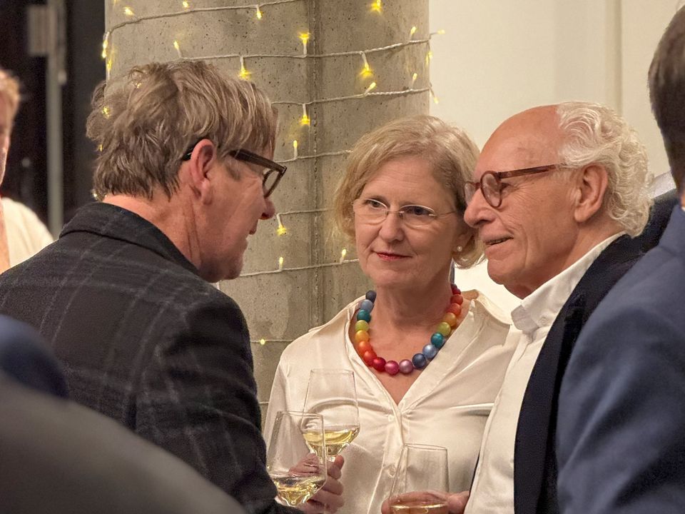 Nahaufnahme eines Gesprächsaustauschs zwischen mehreren Personen bei einem Architektur‑Event, mit Getränken und Lichterkette im Hintergrund.