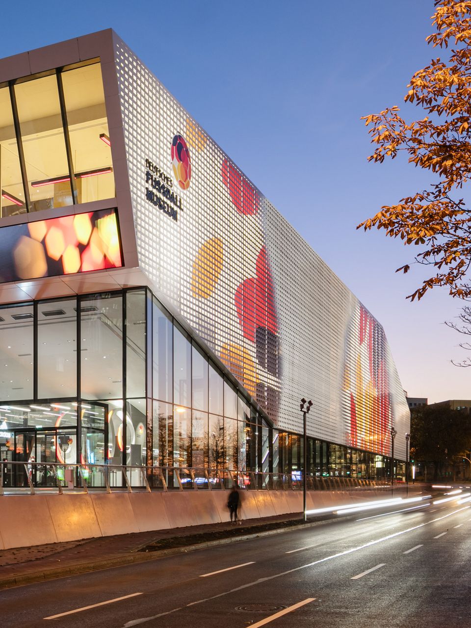 Außenaufnahme des Museums bei Dämmerung mit großformatiger Fassadengrafik, transparentem Eingangsbereich und dynamischen Lichtspuren des Straßenverkehrs.