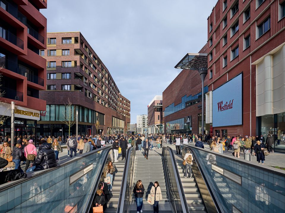 Straßenperspektive eines modernen Einkaufs- und Wohnquartiers mit Rolltreppenabgang, Ziegelfassaden und belebten öffentlichen Bereichen.