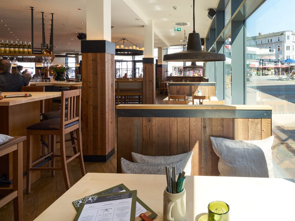 Innenansicht eines Restaurants mit Holzverkleidung, großen Fenstern und gemütlichen Sitzbereichen, moderne und warme Gestaltung