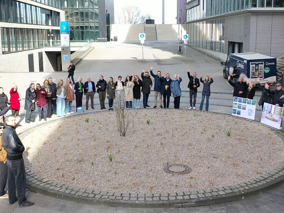 Außenaufnahme einer Gruppe von Personen, die in einem großen Kreis um eine bepflanzte Fläche stehen, umgeben von Bürogebäuden und offenem Stadtraum.