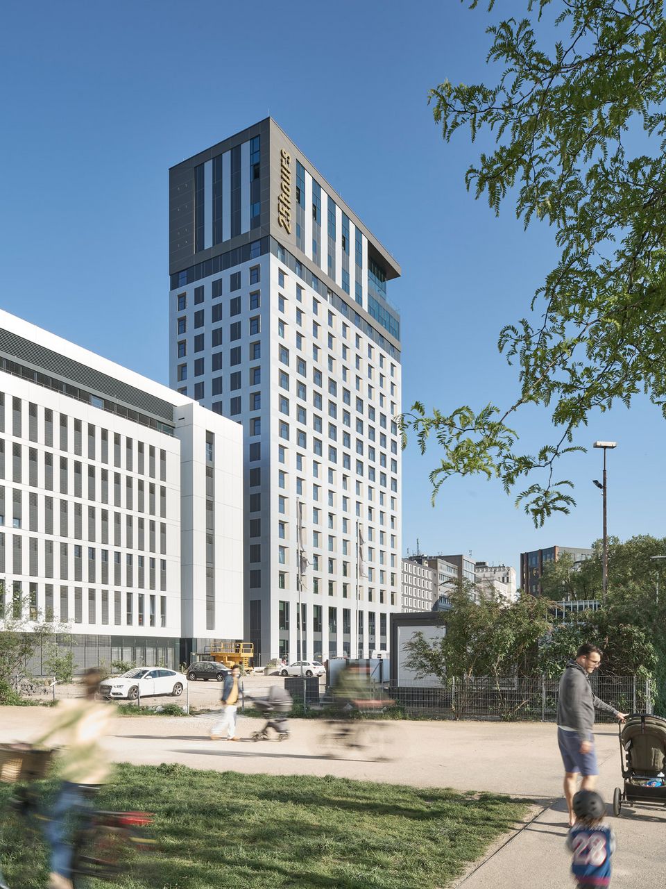 Außenaufnahme eines modernen Hochhauses mit vertikal gerasterter Fassadenstruktur, auskragendem Dachvolumen und angrenzenden Blockrandgebäuden im Stadtquartier.