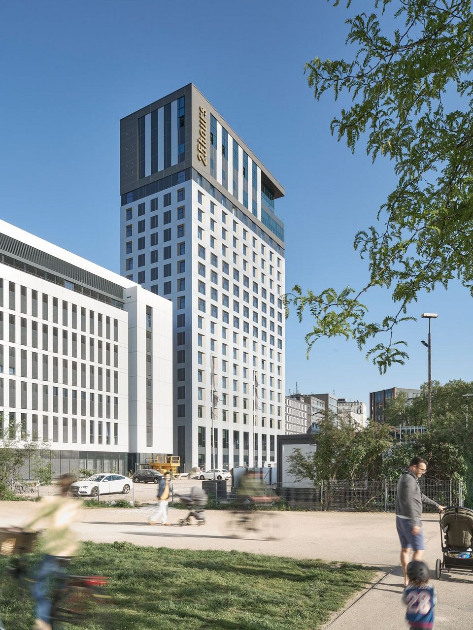 Exterior view of a modern high‑rise building featuring a vertical grid façade, a projecting roof volume and adjacent perimeter‑block structures within the urban district.