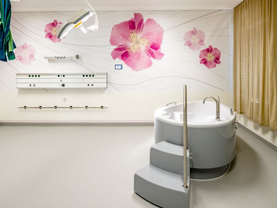 Innenaufnahme eines freundlich gestalteten Geburtsbads mit Rundwanne, floral bedruckter Wand und medizinischer Ausstattung.